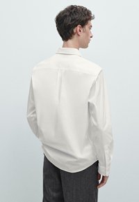 Camicia bianca a maniche lunghe con vestibilità comoda, colletto a punta e una leggera piega nella parte posteriore, abbinata a pantaloni grigi scuri con texture.