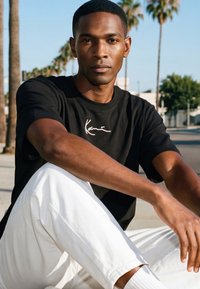 Mann mit kurzen Haaren, der ein schwarzes T-Shirt und weiße Hosen trägt, sitzt im Freien vor Palmen und klarem Himmel.