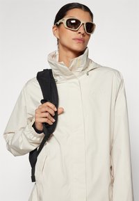 Lys beige vanntett jakke med høy krage og elastiske mansjetter. Svart skulderreim synlig, kombinert med store solbriller.