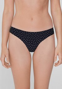 Černé bikini kalhotky s bílými puntíky, nízký pas, hladká textura a elastický pas. Styl s minimálním pokrytím.