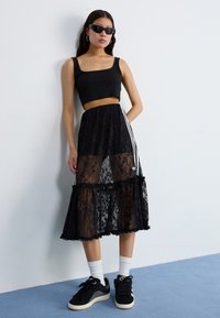 A woman leans against a wall, wearing a black crop top, sheer lace skirt, and black sneakers with white socks. She sports sunglasses.