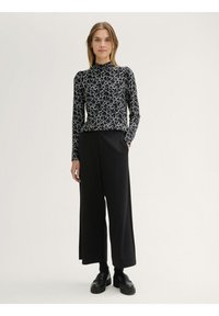 Zwart langemouwen top met wit abstract patroon en hoge hals, gecombineerd met zwarte wijde broek en zwarte platform schoenen.