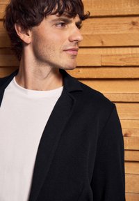 Svart strukturerad blazer över en vit skjorta, med slagkrage och öppen krage, framför en bakgrund av träpanel som skapar kontrast.