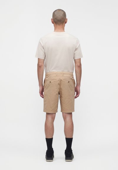 Homme portant un short beige, un t-shirt beige clair, des chaussettes noires et des chaussures noires, debout de dos sur un fond blanc uni.