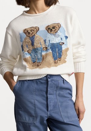 Suéter blanco con dos ositos bordados vestidos con ropa casual, combinado con pantalones azules y manos en los bolsillos.