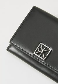 Portofel din piele neagră, cu cusături vizibile și un logo geometric argintiu pe clapeta din față, plasat pe un fundal alb.