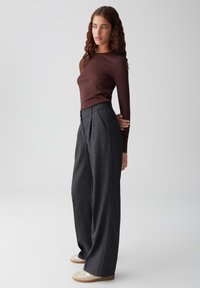 Femme avec des cheveux bruns bouclés portant un top ajusté à manches longues marron, un pantalon large taille haute gris foncé et des baskets beiges.