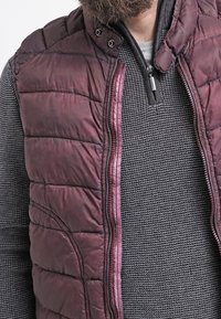 Gilet imbottito bordeaux con zip frontale, dotato di colletto alto e design trapuntato. Sotto, è visibile un maglione grigio scuro con texture.