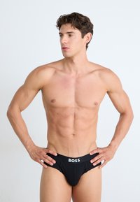 Culotte en coton noir avec un logo blanc "BOSS" sur la ceinture, présentant un tissu lisse et une coupe ajustée. Le modèle pose les mains sur les hanches.