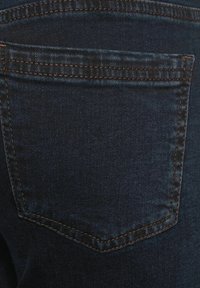 Jean en denim bleu foncé avec une seule poche arrière présentant des coutures visibles et une surface texturée. La poche a un bord supérieur pointu.