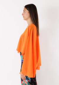 Top leggero in chiffon arancione con un design fluido, maniche lunghe e orlo a strati, abbinato a una gonna a fantasia con dettagli floreali.