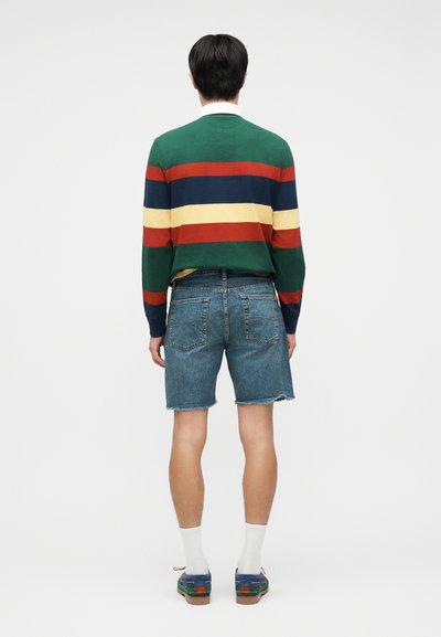 Άνδρας που φοράει πουλόβερ με πράσινες, κόκκινες, μπλε και κίτρινες ρίγες, κοντό τζιν, λευκές κάλτσες και πολύχρωμες μοκασίνες, στέκεται γυρισμένος προς τα πίσω.