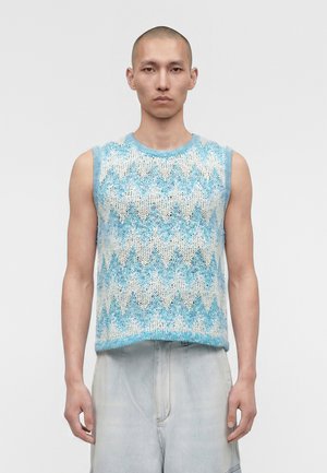 Άνδρας με ξυρισμένο κεφάλι που φοράει αμάνικο πλεκτό γιλέκο με μπλε και λευκά ζιγκ-ζαγκ μοτίβα και φαρδιά ανοιχτόχρωμα denim παντελόνια, στέκεται μπροστά σε απλό φόντο.