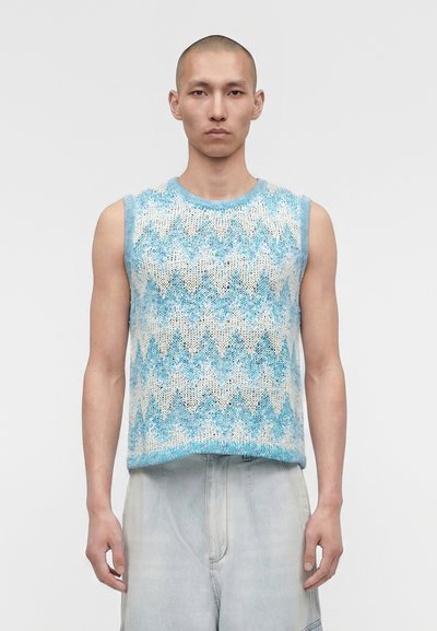 Homme à la tête rasée portant un gilet tricoté sans manches à motif zigzag bleu et blanc et un pantalon en jean léger et ample, debout sur un fond uni.