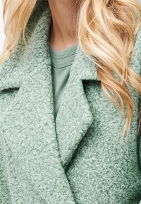 Cappotto verde con una trama densa e pelosa e rever pronunciati, indossato sopra una felpa verde coordinata.