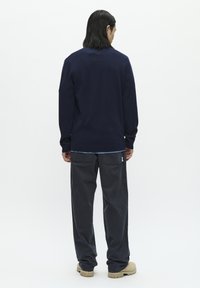 Pulover bleumarin cu manșete și tiv zimțat, asortat cu pantaloni gri închis cu un buzunar aplicat și etichetă cu logo. Fundal deschis.