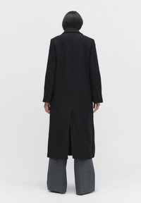 Lång svart kappa med en strukturerad krage, enkel slits baktill och knäppningar vid ärmarna, i kombination med grå vida byxor.