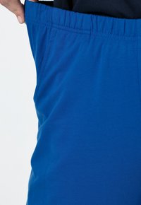 Blå stretchbyxor med elastisk midja, med en slät yta och sidofickor, delvis hållna vid midjan av en hand.