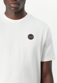 White cotton t-shirt with round neckline. Features a circular embroidered logo on the left chest in black, orange, and blue colors.