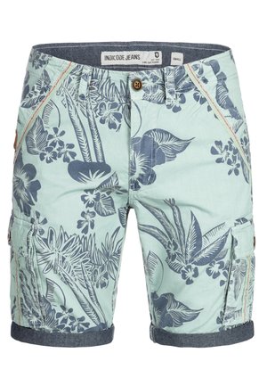 Ung mand stående med hænderne foldet, iført en hvid t-shirt, lyseblå blomstrede cargobukser og hvide sneakers.
