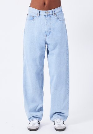 Hellblaue, locker sitzende Jeans aus Denim, getragen mit weißen und grauen Sneakers, von der Taille bis zu den Füßen vor weißem Hintergrund zu sehen.