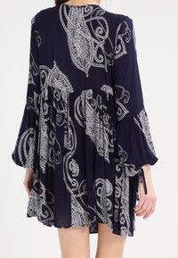 Ein Marinekleid mit einem Paisleymuster, ausgestellten Ärmeln und einer gerafften Taille. Leichter Stoff mit einer strukturierten Oberfläche. Die Länge endet über dem Knie.