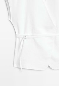 Blouse blanche à manches courtes avec une fine ceinture nouée à la taille et un ourlet arrondi avec une petite fente sur le devant.
