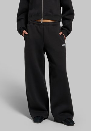 Personne portant un pantalon de survêtement noir à jambes larges et une veste courte zippée assortie, debout les mains dans les poches devant un fond gris.