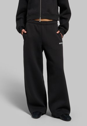 Personne portant un pantalon de survêtement noir à jambes larges et une veste courte zippée assortie, debout les mains dans les poches devant un fond gris.