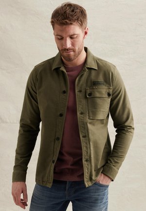 Man draagt een olijfgroene jas met knopen over een bordeauxrode shirt, met één hand in de zak en kijkt naar beneden tegen een effen achtergrond.