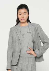 Mulher jovem vestindo um blazer cinza risca de giz, saia plissada a condizer e suéter cinza com detalhes em malha floral, posando com uma mão na anca.