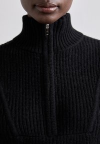 Pull en maille côtelée noire avec col montant et demi-fermeture éclair. Présente une finition texturée et une coupe structurée, mettant en valeur un design minimaliste.