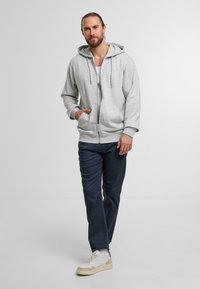 Gray zip-up hoodie with a drawstring hood, worn over a white tank top. Paired with navy trousers and beige-accented sneakers.