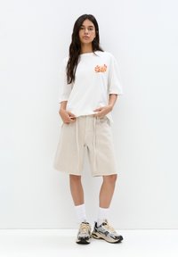 Vit oversized t-shirt med orange grafik, beige löst sittande shorts, vita strumpor och grå sneakers med svarta accenter.