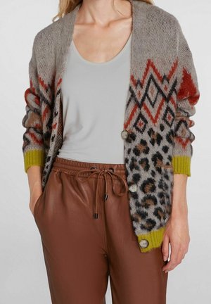 Vrouw draagt een grijze cardigan met geometrische en dierenprints over een wit topje en bruine leren broek met trekkoord.