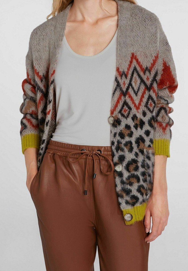 Femme portant un cardigan gris à motifs géométriques et animaliers sur un haut blanc et un pantalon en cuir marron avec cordon de serrage.