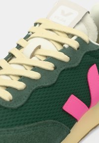 Baskets vertes avec des lacets crème, une étiquette blanche sur la languette portant l'inscription "Veja" et un logo "V" rose sur le côté près de la semelle.