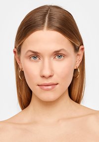 Gouden hoepeloorbellen met een gladde, gepolijste oppervlakte; cirkelvormig; minimalistisch ontwerp; gedragen aan het linkeroor van het model.