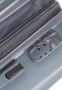 Trolley-Koffer mit einer strukturierten grauen Außenseite, ausgestattet mit einem Zahlenschloss, Reißverschlussfächern und einem stabilen Griff für einfachen Transport.