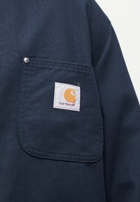 Tmavě modrá látka s obdélníkovou kapsou, na které je přišitá malá bílá nášivka s logem Carhartt v pravém horním rohu.
