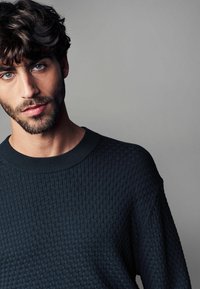 Dunkel strukturierter Pullover mit einem gerippten Rundhalsausschnitt und einem Muster aus kleinen Quadraten, vor einem neutralen Hintergrund.