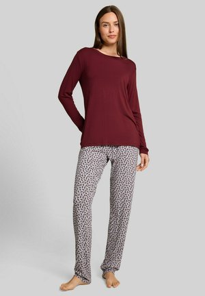 Vrouw staat blootsvoets met een donkerrode top met lange mouwen en lichtgekleurde, patroonpyjamabroek tegen een effen achtergrond.