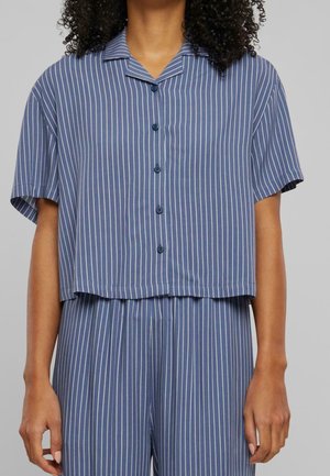 Vrouw in een blauw overhemd met knopen en bijpassende broek met witte verticale strepen, staand voor een effen grijze achtergrond.