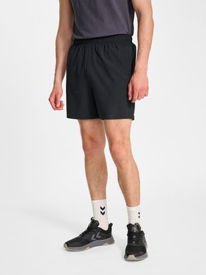 Mand iført sorte atletikshorts, sorte sneakers, hvide sokker med sort chevrondesign og en mørkegrå skjorte, stående på hvid baggrund.
