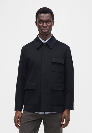 Mann trägt eine schwarze, geknöpfte Jacke mit drei Klappentaschen über einem hellblauen Hemd und steht vor einem einfarbigen weißen Hintergrund.