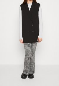Svart väst med dubbelknappning, bärs över en vit långärmad topp, tillsammans med strukturerade grå rutiga byxor och svarta skor.