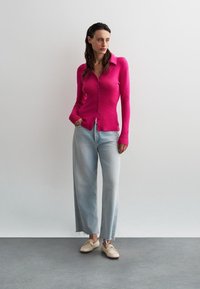 Cardigan lungo fucsia con maniche a lunghe e bottoni, abbinato a jeans larghi azzurri e scarpe beige con texture intrecciata.