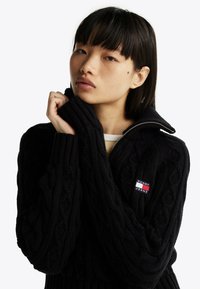 Maglione nero a maglia lavorata con collo alto, cerniera e toppa con logo. Presenta polsini a coste e dettagli testurizzati per una vestibilità aderente.