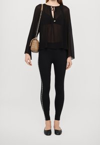 Czarna, prześwitująca bluzka z dekoltem zdobionym kokardą, połączona z czarnymi legginsami z bocznym paskiem z logo oraz płaskimi czarnymi butami. Brązowa, pikowana torebka na ramię.