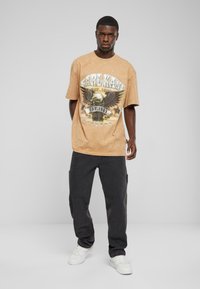 Tan grafisk t-shirt med ørneret design og teksten "Karl Kani 1989." Sammen med mørkegrå cargo bukser og hvide sneakers.